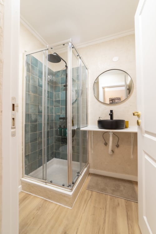 Modern bathroom in Bica Mini Hostel apartment with glass shower, black sink, round mirror, and wooden flooring in Lisboa