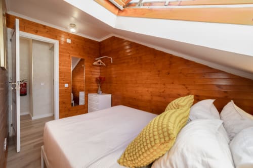 Modern loft bedroom at Bica Mini Hostel in Lisbon with wooden walls, white bedding, yellow pillow, skylight, and ensuite