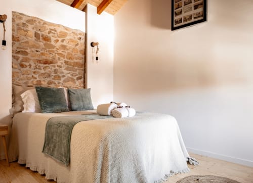 Quarto rústico em Recantos D'Almerinda em Pombal com parede de pedra, roupa de cama creme e vigas de madeira