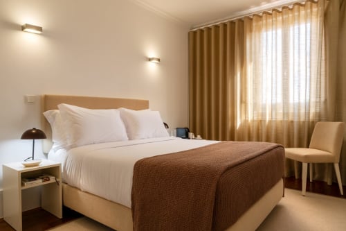 Quarto moderno no Oporto Comfort Charming no Porto com cabeceira bege, roupas de cama brancas, manta marrom e vista para a cidade