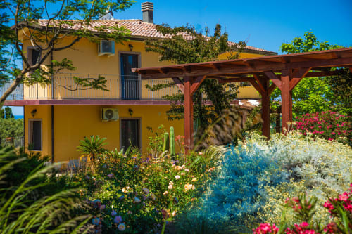 Villa Sardegna Explora a Modolo con facciata gialla, pergolato in legno e giardino mediterraneo colorato sotto cielo azzurro