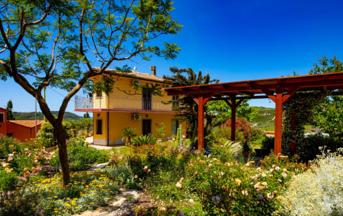 Villa gialla con pergolato rosso circondata da giardino fiorito sotto cielo blu a Modolo, Sardegna Explora House