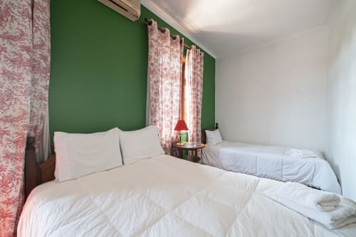 Quarto luminoso da Casa Augusta com camas de solteiro, cortinas florais rosa, parede de destaque verde e roupa de cama branca em Penafiel