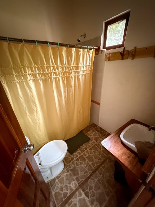 Baño tradicional en el hostal Cayuco Maya de Bacalar con cortina de ducha amarilla, inodoro y lavamanos