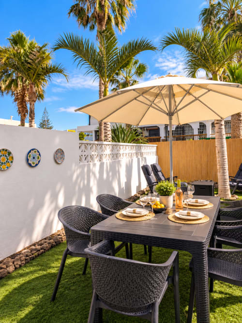 Outdoor dining patio at Manage Home Tenerife with black chairs, umbrella, palm trees, and decorative plates on white wall.