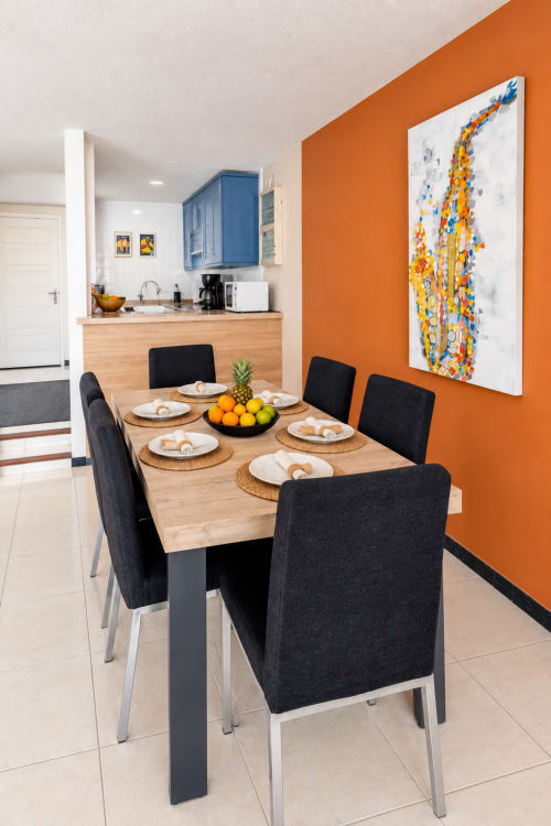 Modern dining area with wooden table, black chairs, orange accent wall, and kitchen in Tenerife apartment