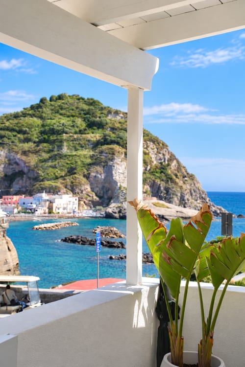 Terrazza costiera a Sant'Angelo con vista sul mare turchese, pergola bianca, pianta in vaso verde e il colorato villaggio adagiato