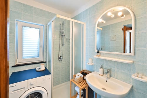 Bagno moderno con piastrelle azzurre, specchio illuminato, box doccia in vetro e lavatrice nell'appartamento Sant'Angelo