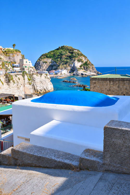 Moderna piscina a sfioro bianca con interno blu che si affaccia sul porto di Sant'Angelo e l'isola rocciosa, Ischia Italia