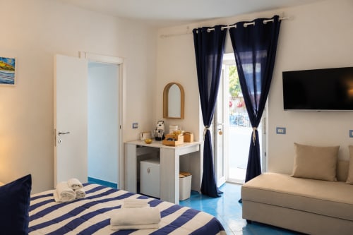 Camera Chiaia di Rose con letto a righe blu, divano beige, tende scure, TV schermo piatto e porta verso la terrazza