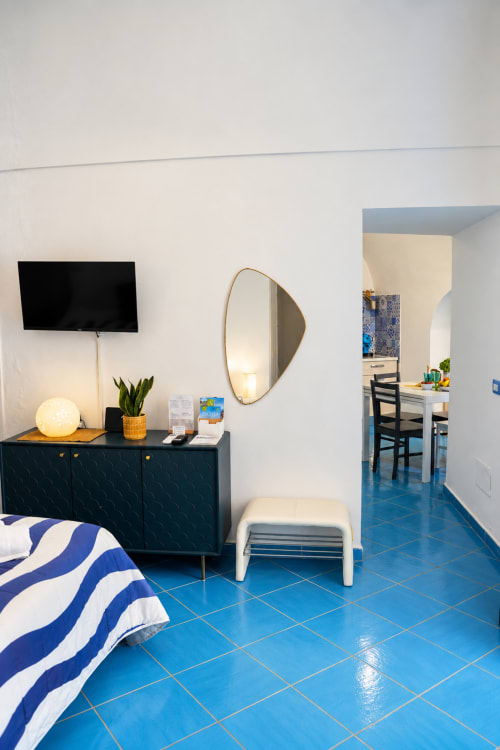 Modern apartment bedroom at Chiaia di Rose with blue tiled floor, dark dresser, round mirror, and doorway to kitchen area