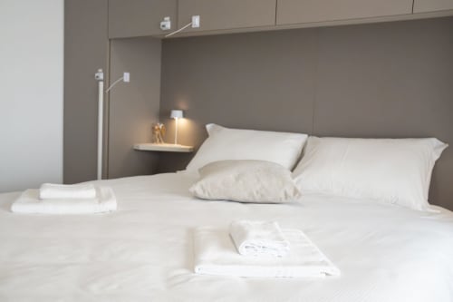 Camera da letto moderna a Ferrara con biancheria bianca, testata grigia, asciugamani piegati e lampada comodino