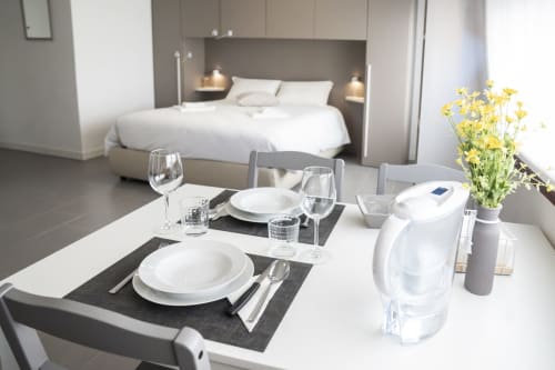 Modern studio apartment a Ferrara con zona pranzo per due persone, letto sullo sfondo, fiori gialli e cucina completamente attrezzata
