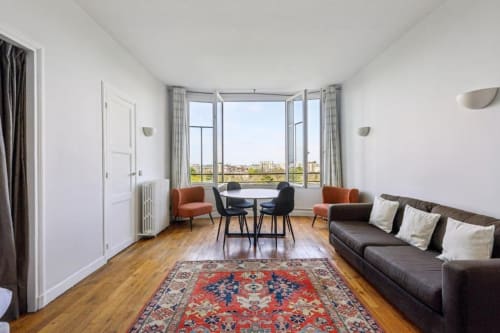 Suite moderne de la Résidence Aurmat avec baies panoramiques, tapis rouge, canapé brun et vue sur la ville de Boulogne-Billancourt
