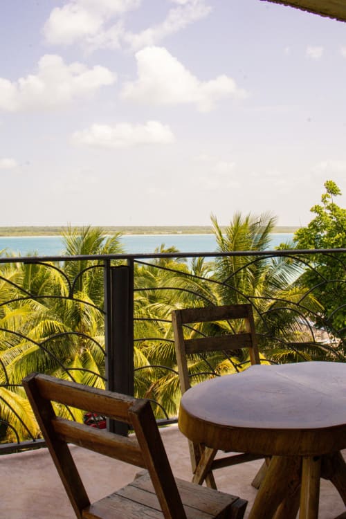 Silla y mesa de madera en la terraza lacustre del hostel Cayuco Maya en Bacalar, México, con vistas al agua turquesa y palmeras