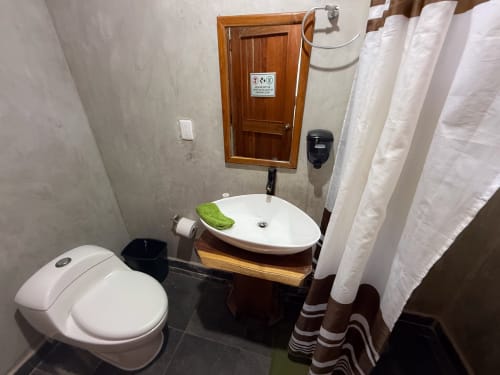Baño moderno del hostal Cayuco Maya en Bacalar con inodoro blanco, lavamanos redondo, espejo de madera y cortina de ducha