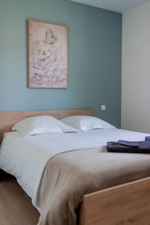 Modern bedroom in Résidence des Sables apartment with teal accent wall, wooden bed frame, white bedding, and Buddha sketch