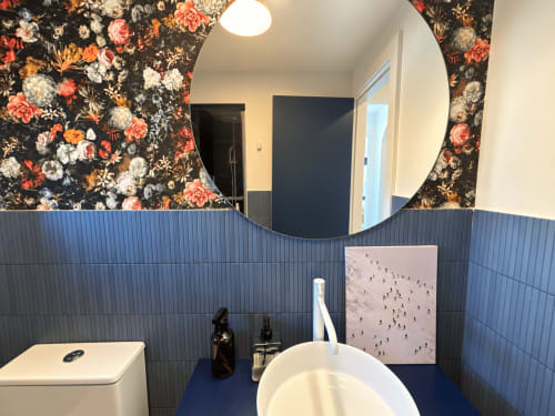 Baño moderno de la casita El Román en Baiona: azulejos azules, espejo redondo, lavamanos blanco y papel tapiz floral