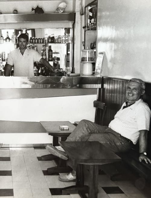 Barman derrière le comptoir du bar de l'Hôtel Thalassa à Cargèse, avec un client souriant assis à une table