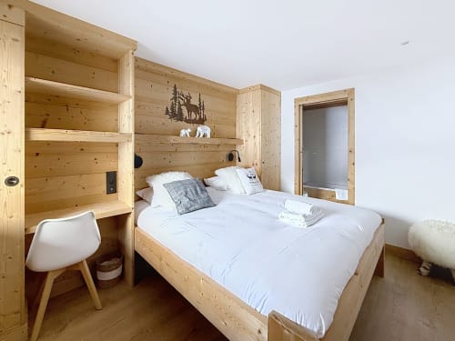 Chambre cozy en pin avec lit queen, literie blanche, étagères en bois décorées de paysages montagnards, fauteuil moderne et accès à la salle de bain