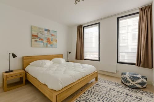 Chambre lumineuse et moderne de l'appartement The Livorno à Bruxelles avec lit en bois, literie blanche, grandes fenêtres et décoration contemporaine