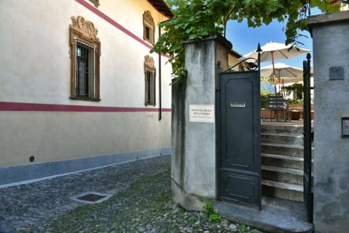 Ingresso con cancellata nera della Roseto del Drago B&B a Ponte In Valtellina, cortile ricoperto di edera e ombrelloni