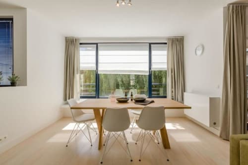 Élégante salle à manger d'appartement bruxellois avec table en bois, chaises claires et baies vitrées donnant sur la verdure