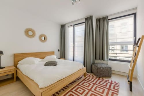 Chambre moderne de l'appartement The Livorno Brussels avec lit en bois, literie blanche, rideaux vert sauge et tapis géométrique rouge