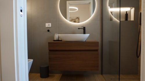 Bagno moderno di Marelia Suites Vasto con specchio rotondo illuminato, lavabo bianco sospeso, mobile in legno e doccia aperta