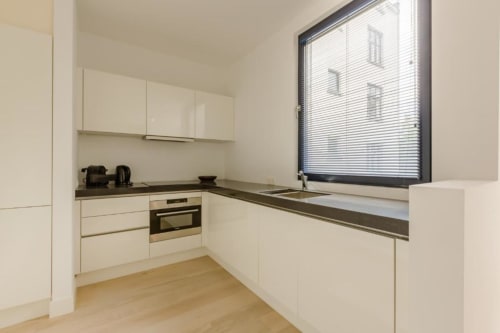 Cuisine blanche moderne dans un appartement bruxellois avec plan de travail sombre, four encastré et grande fenêtre