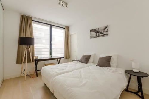 Appartement moderne à Bruxelles au Livorno avec deux lits blancs, bureau en bois et grandes fenêtres aux rideaux beige