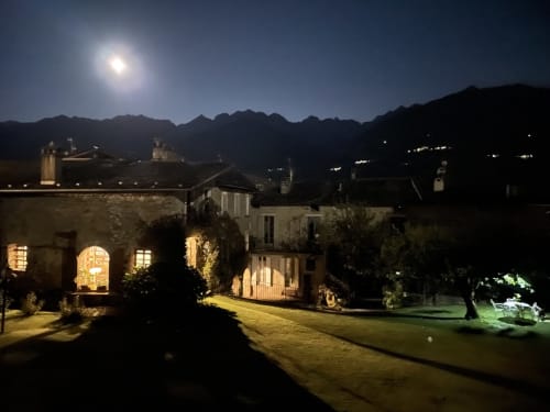 Cortile del Roseto del Drago illuminato dalla luna con edifici in pietra e lo sfondo delle montagne a Ponte in Valtellina
