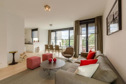 Élégant salon d'un aparthotel bruxellois avec canapé gris, coussins rouges, table ronde et vue sur balcon à The Livorno