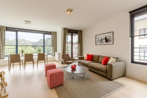 Appartement moderne et élégant à Bruxelles avec canapé beige, accents rouges et baies vitrées donnant sur la verdure.
