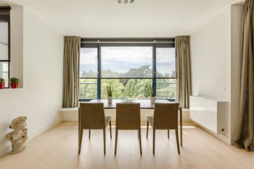 Appartement moderne à Bruxelles avec espace repas, trois chaises, grandes fenêtres donnant sur la verdure, rideaux beige