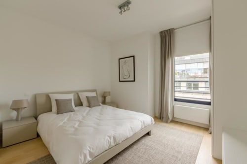 Chambre moderne de l'appartement The Livorno Brussels avec lit capitonné beige, literie blanche et baies vitrées du sol au plafond