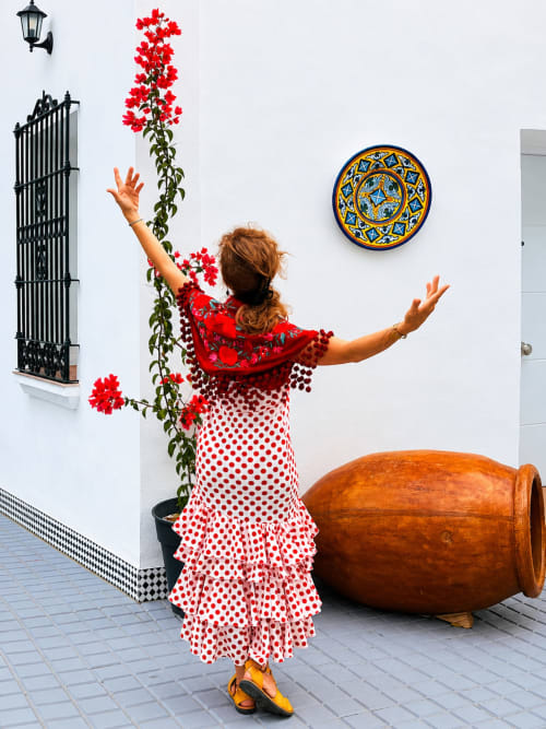 Mujer con vestido rojo de lunares y brazos levantados en el hotel Alcazaba Salobreña, rodeada de flores rojas vibrantes