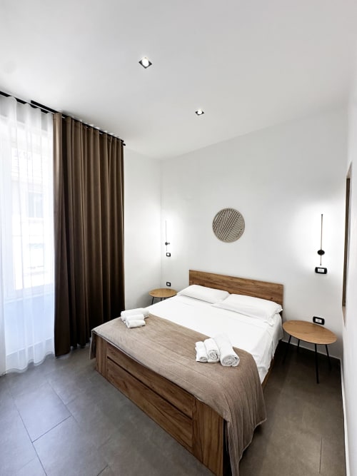 Camera matrimoniale moderna presso I colori di Napoli B&B a Napoli con letto in legno, biancheria bianca e vista sulla città attraverso finestre con cornici marroni