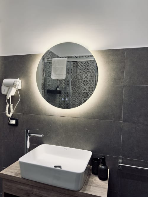 Moderno bagno presso I colori di Napoli con specchio illuminato circolare, lavabo bianco a bacinella e doccia con piastrelle decorative