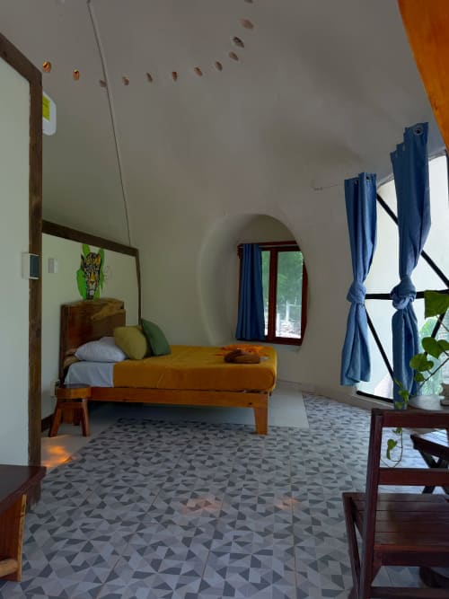 Habitación con bóveda geodésica en el hostal Cayuco Maya de Bacalar, con cama de madera, cortinas azules y piso de azulejos geométricos