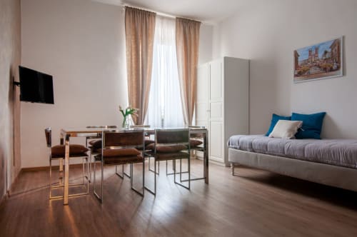 Modern apartment in Roma with wooden floors, dining table, bed with blue pillows, TV, and large windows with beige curtains