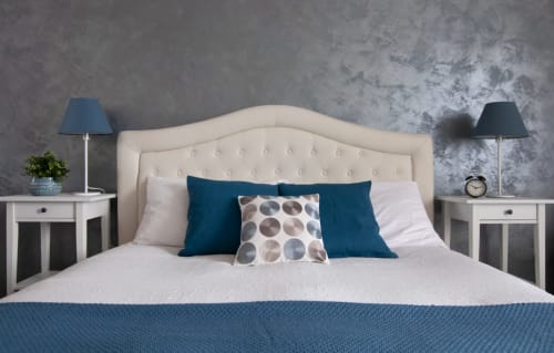 Modern bedroom at UrbanFlat Roma with cream headboard, teal pillows, white bedside tables, and grey textured wall backdrop.