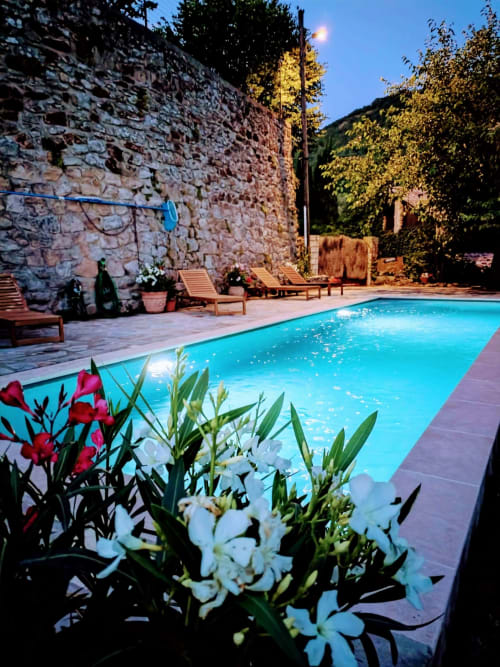 Illuminated pool at twilight with red and white flowers, stone building, lounge chairs, and mountain views in South France.
