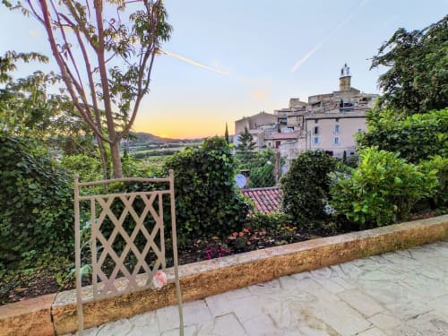 Terrace with lattice gate overlooking Venterol village at sunset, enclosed garden with flowering plants and mountain views.