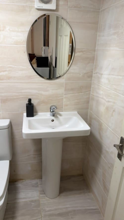 Modern bathroom with white pedestal sink, round mirror, and beige tile walls in England Furnished Homes