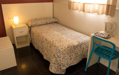 Habitación individual en el Hostal San Vicente II de Sevilla con cama individual, mesilla de noche, lámpara, silla azul y ventana con