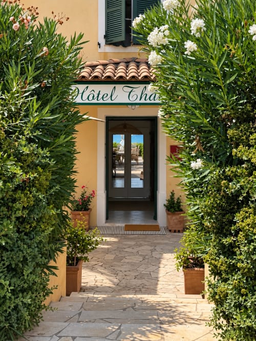 Entrée de l'Hôtel Thalassa à Cargèse avec plantes fleuries et pots en terre cuite encadrant les portes noires