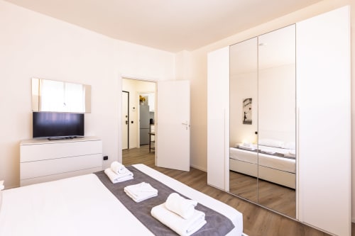 Solumnia Urban Nest, camera da letto in Milano con armadio a specchio, TV e biancheria bianca su tappeto grigio