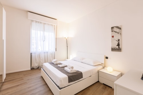 Camera moderna presso Solumnia Urban Nest a Milano con letto matrimoniale, comodino bianco, lampada da terra e finestre con vista sulla città
