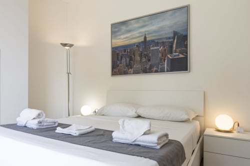 Camera da letto moderna a Milano con vista sullo skyline, letto bianco, asciugamani freschi, lampada da terra e lampada da comodino presso Solumnia